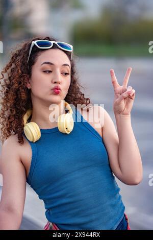 Verspielte junge Frau mit lockigen Haaren, die ein Friedenszeichen und knisternden Lippen machen, Kopfhörer und Sonnenbrille im Stadtpark trägt. Stockfoto