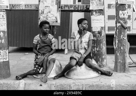 Zwei junge Frauen in Ghana sitzen auf großen Schüsseln, eine mit einem Handy, die einen Moment der Ruhe und der modernen Kommunikation während der täglichen Arbeit darstellen. Stockfoto