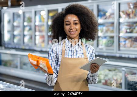 Ein lächelnder Mitarbeiter im Lebensmittelgeschäft hält ein Tablet und ein Produkt in der Tiefkühlhalle und zeigt den Kundenservice und die Technologienutzung im Einzelhandel. Stockfoto