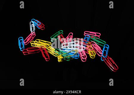 Eine einzelne gelbe Sicherheitsnadel zusammen mit mehreren farbigen Büroklammern auf schwarzer Oberfläche. Horizontales Foto mit Kopierraum. Stockfoto