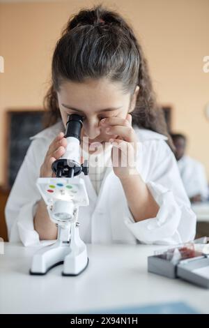 Vertikale mittelgroße Großaufnahme eines Mädchen aus dem Nahen Osten mit Labormantel, das während des Chemieunterrichts am Schreibtisch im Klassenzimmer mit Mikroskop sitzt Stockfoto