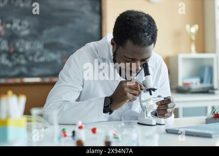 Junger afroamerikanischer Chemielehrer mit weißem Labormantel, der am Schreibtisch im Klassenzimmer mit dem Mikroskop sitzt Stockfoto
