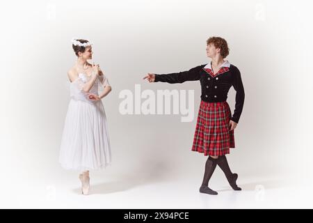 Romantische Szene. Ballerina in fließendem Kleid und Blumenkrone interagiert anmutig mit ihrem Partner in traditioneller schottischer Kleidung gegen weißes Studio Stockfoto