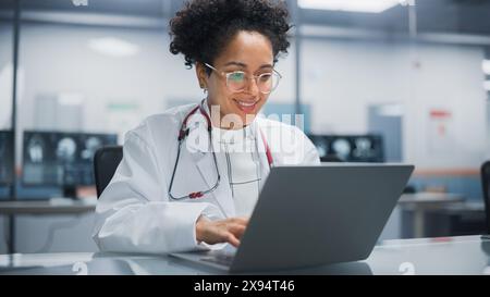 Krankenhausarztpraxis: Porträt junger vielversprechender Schwarzer Arzt arbeitet auf Laptop-Computer. Weibliche Ärztin im weißen Labormantel auf der Suche nach Patientenbehandlung während sie an ihrem Schreibtisch sitzt Stockfoto