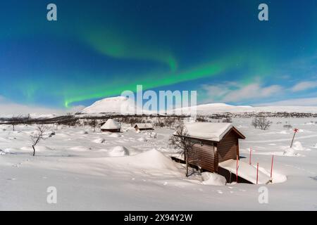 Grüne Nordlichter (Aurora Borealis) über typischen Holzhütten in der schneebedeckten Landschaft, beleuchtet von Vollmond, Finnland Stockfoto