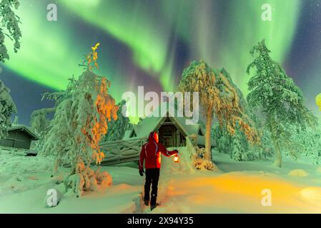 Mann mit Laterne vor einer typischen finnischen Hütte (kota) im schneebedeckten Wald unter Nordlichtern (Aurora Borealis), Pallas-Yllas Stockfoto