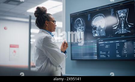 Medical Science Hospital: Selbstbewusste schwarze Neurologin, Neurowissenschaftlerin, Neurochirurg, schaut auf TV-Bildschirm mit MRT-Scan mit Gehirnbildern, denkt über die Behandlungsmethode für kranke Patienten nach. Leben Retten Stockfoto