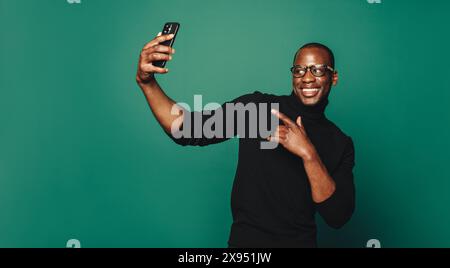 Sorgloser, lächelnder Mann in lässiger Kleidung macht ein Selfie mit einem Smartphone auf grünem Hintergrund. Er posiert selbstbewusst und zeigt seine Mode. Stockfoto