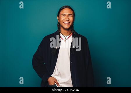 Selbstbewusst und fröhlich steht ein junger Mann mit langen geflochtenen Haaren stilvoll vor blauem Hintergrund in legeren Kleidern und Schmuck. Stockfoto