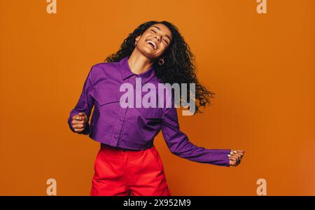 Fröhliche Frau mit lockigen Haaren tanzt fröhlich auf einem leuchtend orangen Hintergrund und präsentiert ihr stilvolles und modisches Outfit. Stockfoto