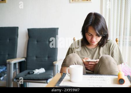 Traurige Frau sitzt am Tisch im Zimmer mit Smartphone und trinkt Tee Stockfoto