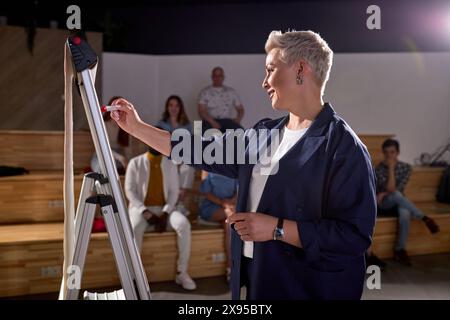 FemaleCollege Professor hält Vortrag im Klassenzimmer voller multiethnischer Studenten. Der Talentierte Sprecher Zieht Die Aufmerksamkeit Des Publikums Auf Sich, Schreibt An Bord, Spricht Stockfoto
