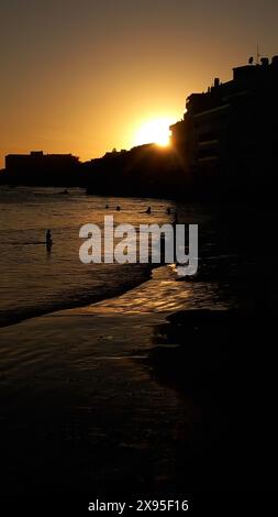 Silhouetten von Menschen, die den Strand bei Sonnenuntergang genießen, während die goldene Sonne hinter Gebäuden untergeht und ein warmes Licht ausstrahlt Stockfoto