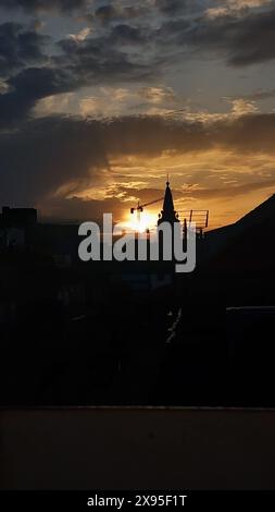 Sonnenuntergang über einer Stadtlandschaft mit der Silhouette eines Uhrenturms und Baukränen vor einem dramatisch bewölkten Himmel Stockfoto