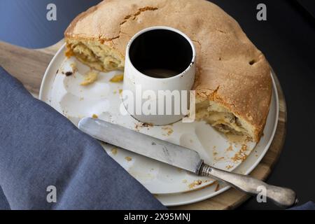 Blick von oben auf einen Apfelschwammkuchen in einer weißen abnehmbaren Dose mit Vintage-Messer auf schwarzem Hintergrund mit dunkelblauen Vorhängen in natürlichem Licht Stockfoto