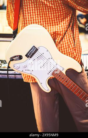 Ein Musiker in einem orangefarbenen karierten Hemd spielt eine weiße E-Gitarre und tritt möglicherweise bei einem Konzert im Freien auf Stockfoto