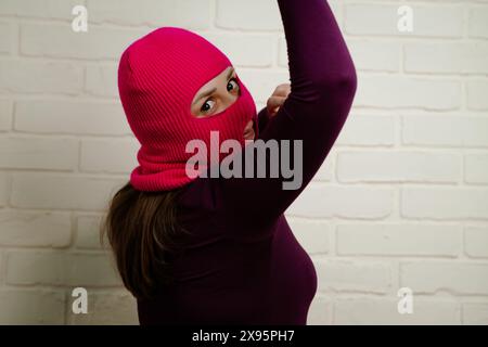 Eine Frau in einer knallrosa Sturmhaube tanzt energetisch und zeigt einzigartige Posen mit ihren Händen vor einem sauberen, weißen Backsteinmauerhintergrund. Stockfoto