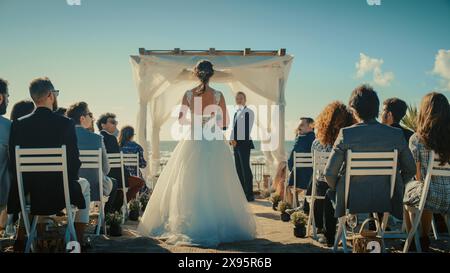Wunderschöne Braut im wunderschönen weißen Hochzeitskleid, die den Gang hinuntergeht, während der Bräutigam an einem Zeremonie-Ort im Freien in der Nähe des Meeres mit fröhlichen multiethnischen und vielfältigen Freunden wartet. Stockfoto