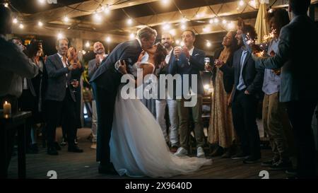 Schöne Braut in weißem Kleid und Bräutigam im stilvollen schwarzen Anzug feiern die Hochzeit auf einer Abendfeier. Jungvermählte tanzen an einem Veranstaltungsort mit den besten multiethnischen Freunden. Stockfoto
