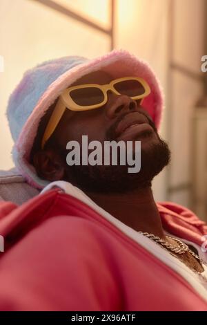 Vertikale Nahaufnahme eines jungen Schwarzen Mannes mit funky rosa Outfit und Sonnenbrille, der sich drinnen in dunklem Licht auf der Couch entspannt Stockfoto