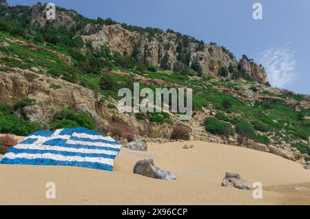 Ein riesiger Stein mit gemalter griechischer Flagge an einer Sandküste mit bewaldeten Bergen Stockfoto