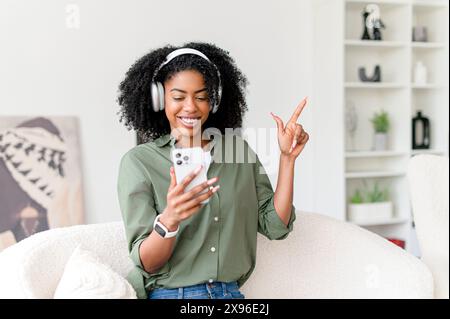 Eine strahlende afroamerikanische Frau genießt ihre Musik, trägt weiße Kopfhörer und hält ein Smartphone, und ihre Freude ist in ihrem komfortablen Zuhause spürbar. Sie verkörpert Freizeit und Konnektivität Stockfoto