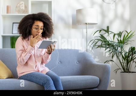 Eine Frau auf einem Sofa reagiert überraschend, während sie sich ein schauriges Video auf einem Tablet in einem gemütlichen Wohnzimmer ansieht. Stockfoto