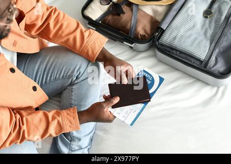 Nahaufnahme von oben: Nicht erkennbarer Schwarzer Mann mit Pass und Tickets, der auf dem Bett im Hotelzimmer sitzt und sich auf die Reise vorbereitet Stockfoto