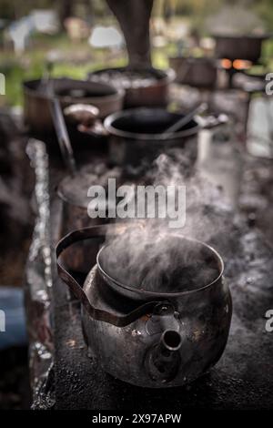 Dampfendes heißes Getränk in einer Teekanne mit alten Metalltöpfen im Hintergrund, die draußen kochen Stockfoto