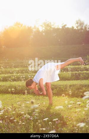 Frau mittleren Alters beim Yoga, die Standing Splits, Urdhva Prasarita Eka Padasana, in einer blühenden Wiese in einem Park während Sonnenuntergang, Deutschland Stockfoto