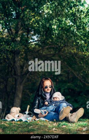 Eine Frau, die in einem Park sitzt, mit ihrem Baby auf dem Schoß, umgeben von Bäumen und Sonnenlicht mit einem Picknick-Set neben ihr Stockfoto