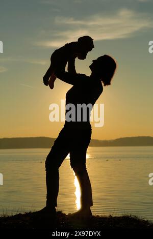 Silhouette eines Elternteils, der ein Kind vor dem Hintergrund eines Sonnenuntergangs am See hebt Stockfoto