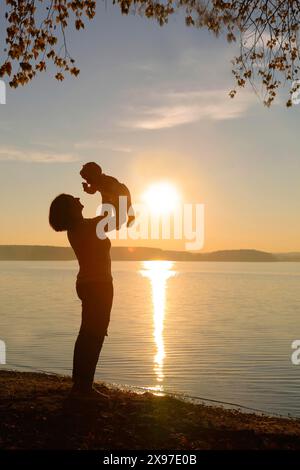 Eine Silhouette eines Elternteils, der während des Sonnenuntergangs ein Kind in der Nähe eines Sees hebt und sich auf dem ruhigen Wasser reflektiert Stockfoto