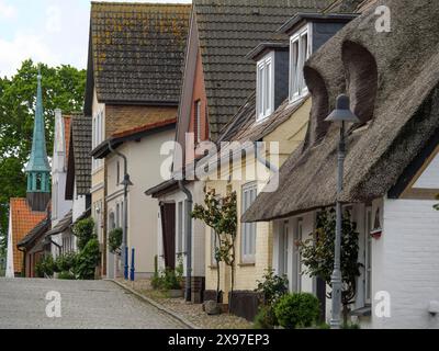 Traditionelle Häuser mit Strohdächern und einem spitzen Turm am Ende der Straße, entlang einer ruhigen, gepflasterten Straße in einem europäischen Dorf, Straße mit Stockfoto