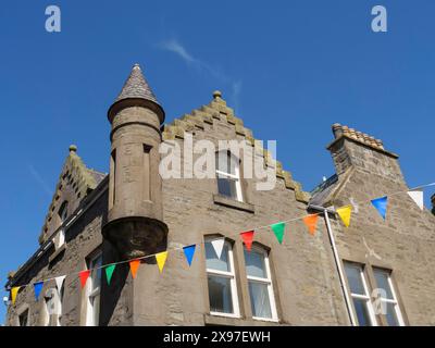 Farbenfrohe Fahnen hängen um ein traditionelles Gebäude mit einem kleinen Turm und Fenstern unter blauem Himmel, graue Häuser mit bunten Fahnen am Meer Stockfoto