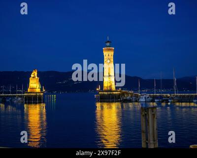 Beleuchteter Leuchtturm und Löwenstatue bei Nacht, deren Lichter im Wasser des Hafens reflektiert werden, türmt sich an einem Hafen auf dem See mit Segeln Stockfoto