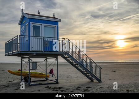 Ein blau-weißer Rettungsturm mit einem gesicherten Boot daneben, vor einem idyllischen Sonnenuntergang am Strand, Sommerabend am Strand mit grüner Düne Stockfoto