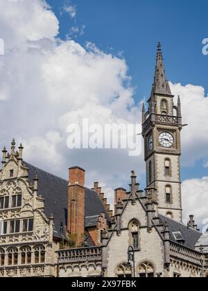 Uhrenturm und gotische Gebäude mit kunstvollen Fassaden unter blauem Himmel mit Wolken, Skyline einer historischen Stadt am Fluss mit alten Fassaden und Stockfoto