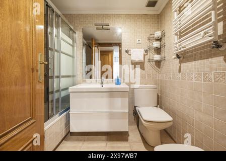 Bad mit Eichenholztür, weißen Porzellantoiletten, hängenden Möbeln mit weißen Schubladen und rahmenlosem Spiegel an der Wand Stockfoto