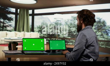 Geschäftsmann mit Greenscreen-Displays auf Geräten, sitzt an seinem Schreibtisch in einer luxuriösen Villa im Wald. Ein reicher Unternehmer, der auf Bildschirmen mit isoliertem Copyspace blickt, arbeitet auf einem Laptop und Tablet. Stockfoto