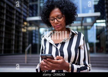 Erwachsene afroamerikanische Frau in formellem Anzug, die vor dem Bürogebäude mit Mobiltelefon steht Stockfoto