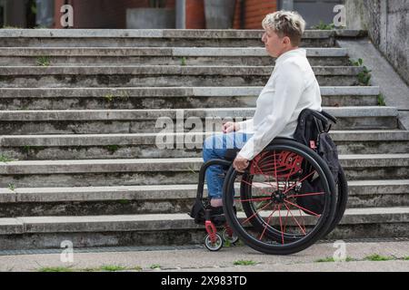 Frau im Rollstuhl hält an und wartet unten auf einer unzugänglichen Straßentreppe, die nicht hinaufgehen kann. Schwierigkeiten- und Behindertenkonzepte. Stockfoto