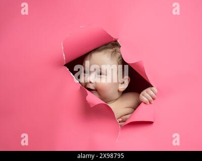 Der süße kaukasische neugeborene Junge blickt aus einem Loch in einem papierrosa Hintergrund. Stockfoto