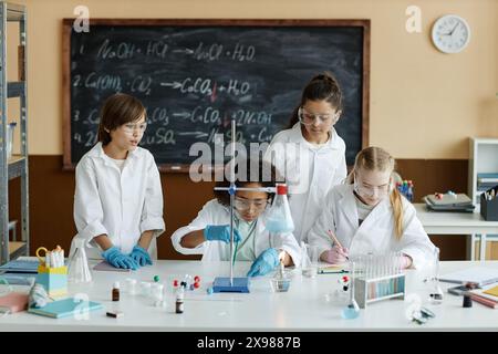 Gruppe von Mittelschüler mit Laborkittel und Schutzbrille, die im Unterricht am Tisch sitzen und stehen, um Experimente im Chemiekurs zu machen Stockfoto