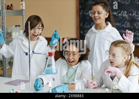 Multiethnische Gruppe glücklicher Jungen und Mädchen in Labormänteln und Schutzbrillen, die chemische Reaktionen in konischen Flaschen untersuchen Stockfoto