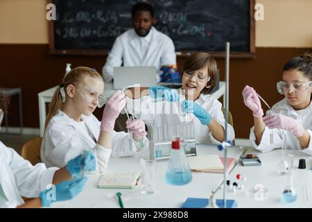 Hochwinkelansicht von ethnisch unterschiedlichen Kindern, die am Tisch sitzen und Laborexperimente in der modernen Chemieklasse machen, während ihr Lehrer an einem Laptop arbeitet Stockfoto