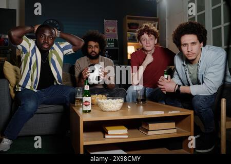 Gruppe ethnisch unterschiedlicher männlicher Freunde, die spät abends im Wohnzimmer sitzen und Fußballspiele im Fernsehen anschauen Stockfoto