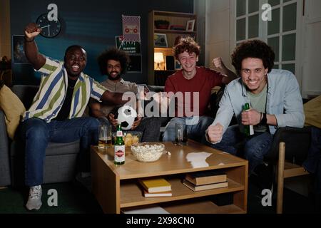 Multiethnische Gruppe fröhlicher junger Männer, die im Wohnzimmer sitzen und sich Fußballspiele im Fernsehen ansehen Stockfoto