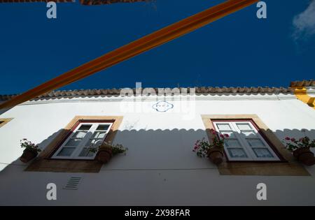 Rua Direita, Óbidos, Portugal. Blick auf den zweiten Stock der traditionellen weiß getünchten Häuser auf der Kopfsteinpflasterstraße, Rua Direita. Bunte Stoffbänder im Zickzack zwischen den Gebäuden über den zahlreichen Boutiquen, Geschäften und Restaurants, die die enge Straße aus dem 13. Jahrhundert im Erdgeschoss säumen. Stockfoto