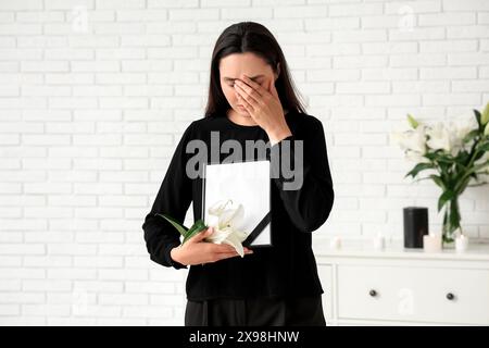 Junge weinende Frau mit Trauerlilienblume und Bilderrahmen im Zimmer Stockfoto